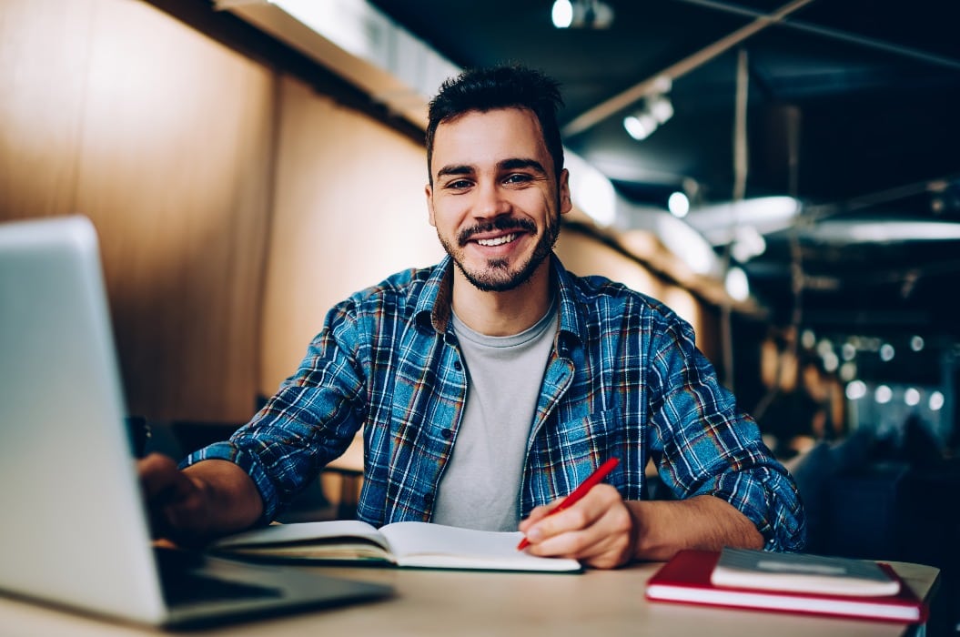 Portrait of cheerful male student enjoying learning in coworking office using laptop computer for research,happy freelancer looking at camera during making project for remote job making notes.